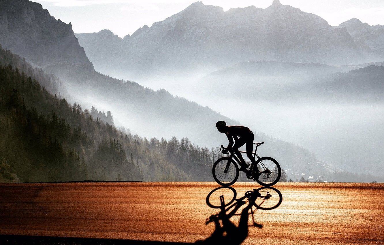 A road cyclist silhouetted against misty alpine peaks, riding a sunlit mountain road
